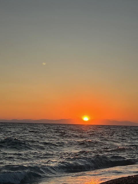 The sun low over a calm sea at the horizon, distant hills in the golden haze.